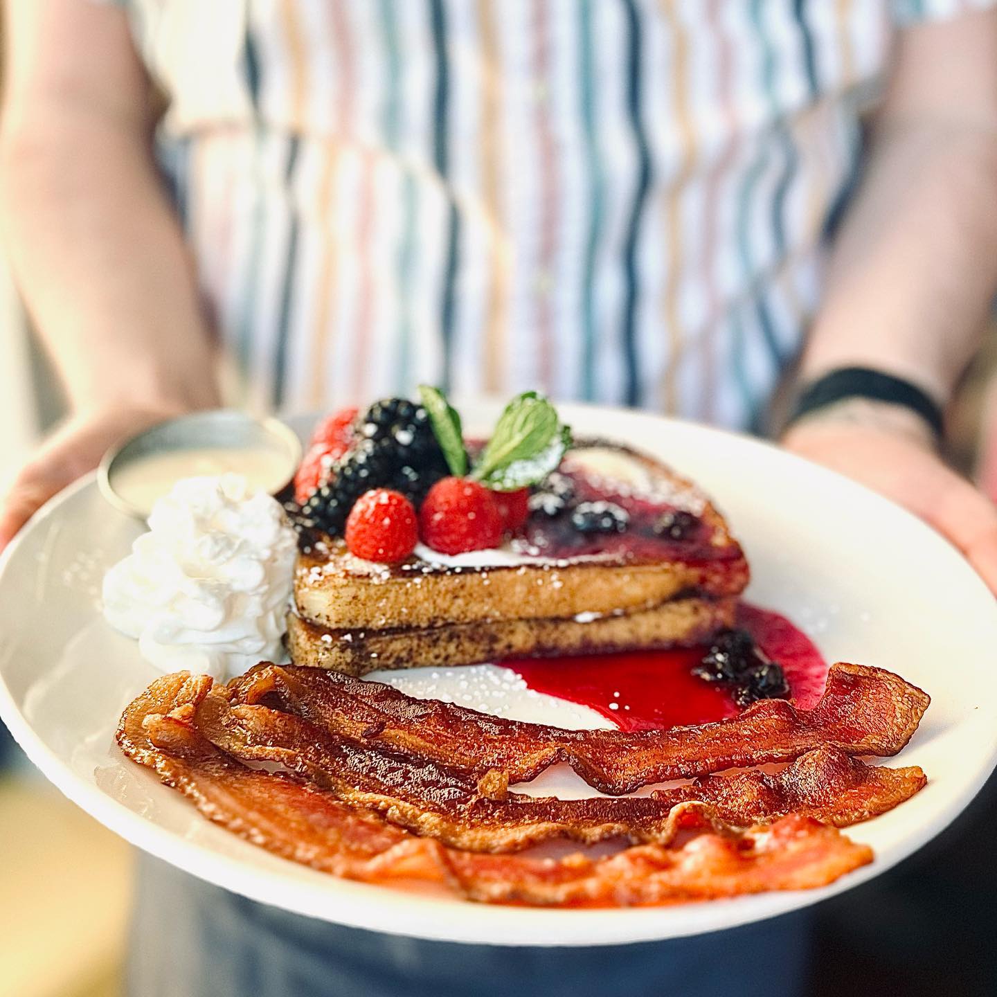 Person holding a plate of french toast, bacon, and fruit at Aurora